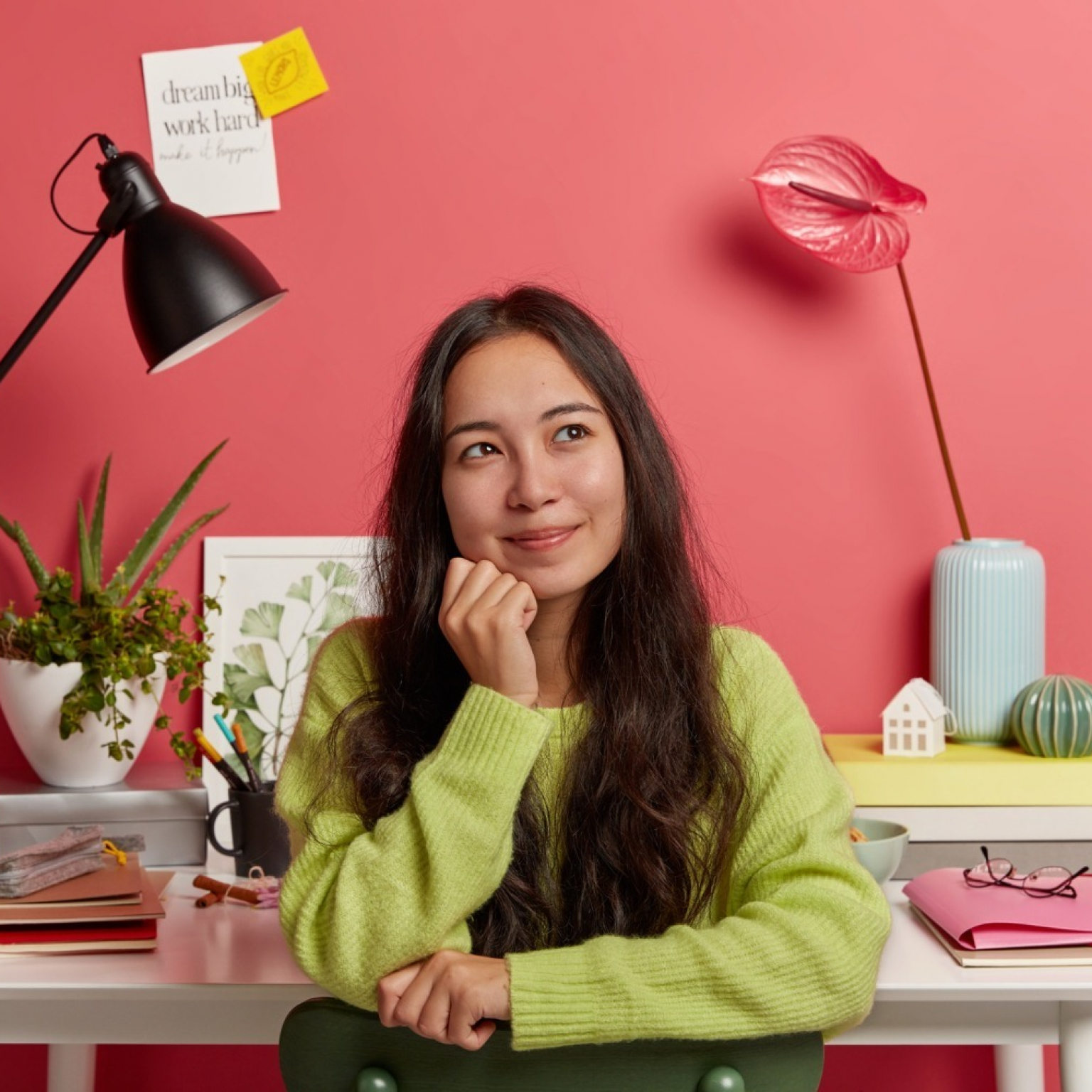 pleased-asian-girl-sits-against-desktop-with-all-necessary-supplies-studying-smiles-daydreams-during-break-poses-chair-against-bright-rosy-wall-sticky-notes-with-inscription