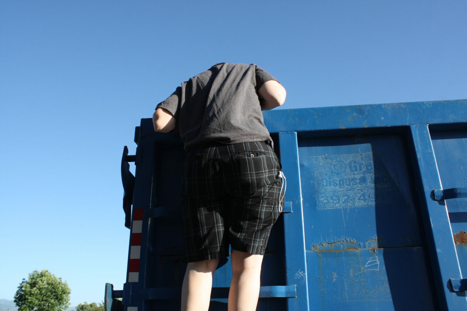 low angle view of a man climbing a blue Junk It dumpster bin doing an unsafety dumpster practices