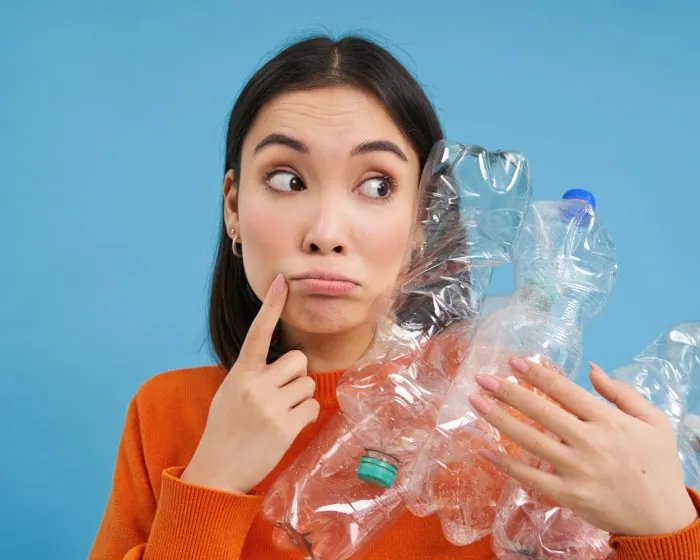 Asian woman holding plastic bottles while thoughtfully considering recycling options - emphasizing the importance of responsible waste management and garbage dumpster rental in Hamilton