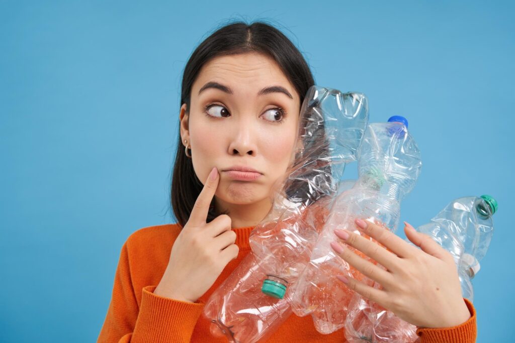 Thoughtful Asian woman holding plastic bottles - looking aside and considering the importance of recycling - while also thinking about efficient garbage bin rental in Toronto for waste management