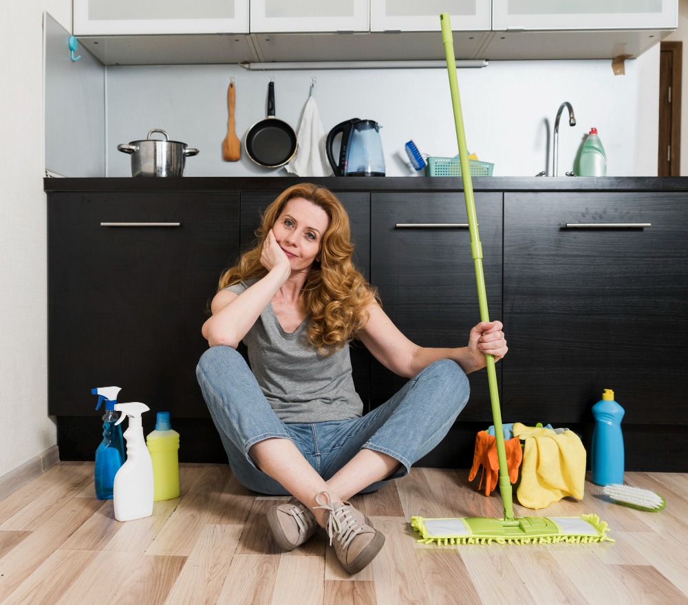 A woman sitting on the floor holding a green mop-appearing thoughtful-with the topic of FAQs on junk bin rental in Barrie in mind