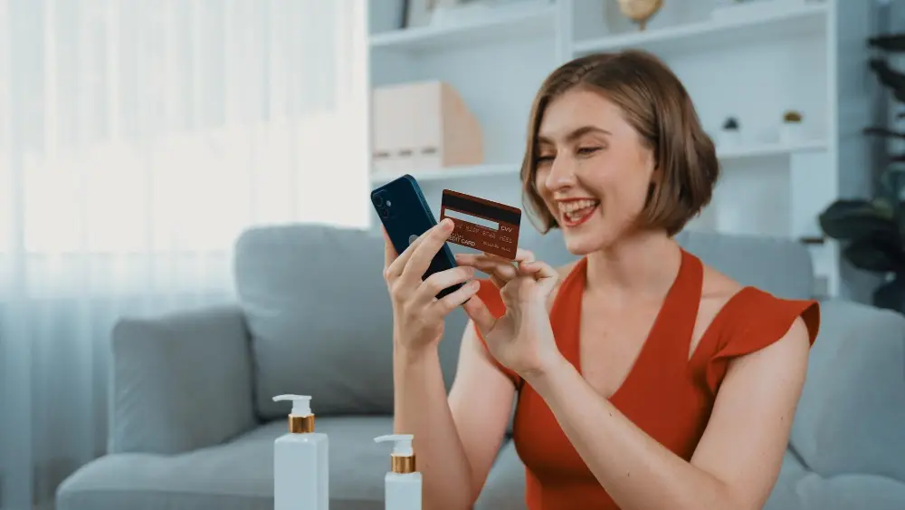A woman in a red shirt smiling as she holds her phone and card, ready to book hassle-free bin rental in Edmonton for her waste disposal needs