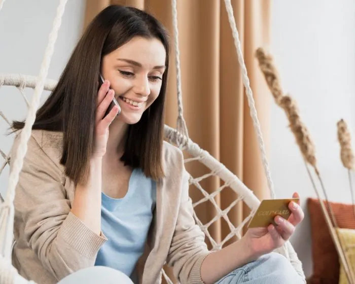 A brown-haired woman on the phone, smiling at her gold card, possibly considering garbage bin rental in London, Ontario for her upcoming decluttering project